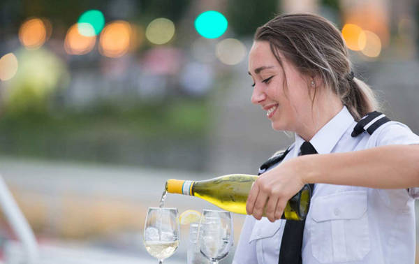 A smiling waitress in uniform pours white wine into glasses, with a blurred background of lights and greenery.