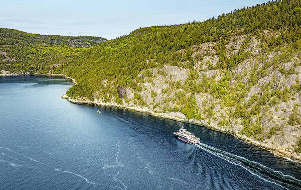 Aerial view of a ferry cruising through a winding, forested river. The vessel leaves a trail in the deep blue water, with rocky cliffs and dense green trees lining the riverbanks under a clear sky.