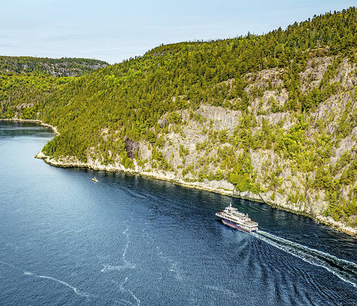 Aerial view of a ferry cruising through a winding, forested river. The vessel leaves a trail in the deep blue water, with rocky cliffs and dense green trees lining the riverbanks under a clear sky.