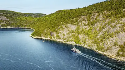 Aerial view of a ferry cruising through a winding, forested river. The vessel leaves a trail in the deep blue water, with rocky cliffs and dense green trees lining the riverbanks under a clear sky.