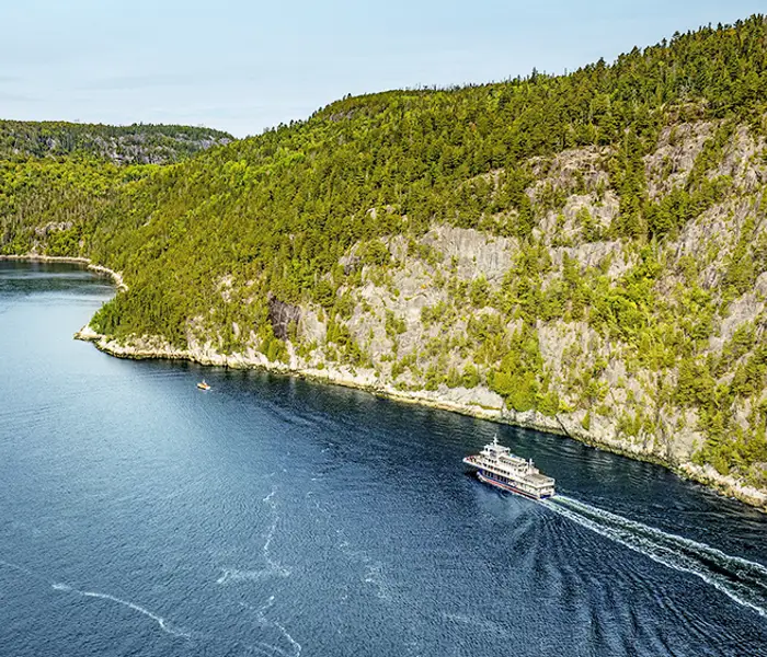 Aerial view of a ferry cruising through a winding, forested river. The vessel leaves a trail in the deep blue water, with rocky cliffs and dense green trees lining the riverbanks under a clear sky.