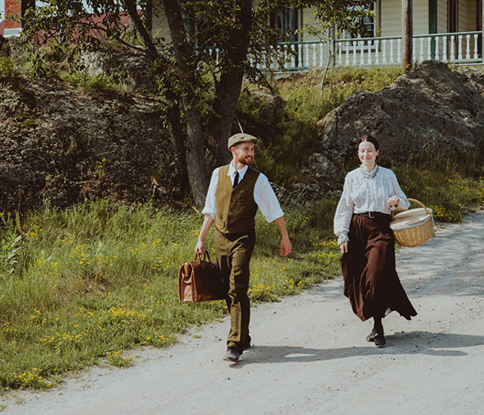 Two characters dressed in period costume are walking on Grosse Île. They are descending a small slope, with a historic house in the background