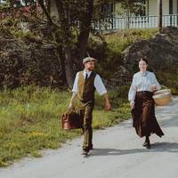 Two characters dressed in period costume are walking on Grosse Île. They are descending a small slope, with a historic house in the background.