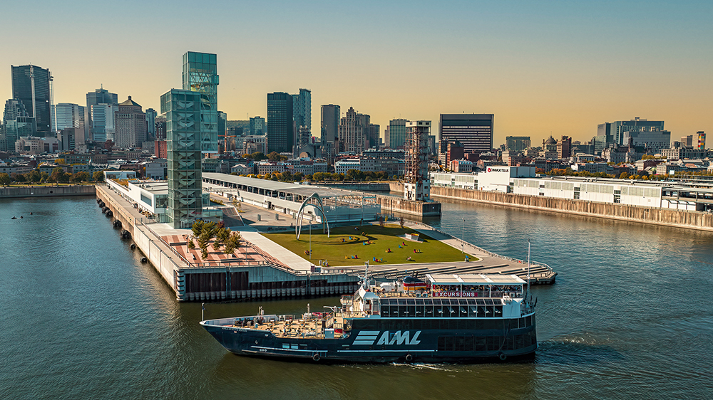 A blue cruise ship with the "AML" logo sails near a modern quay with green spaces, surrounded by the skyscrapers of a city under a clear and sunny sky.