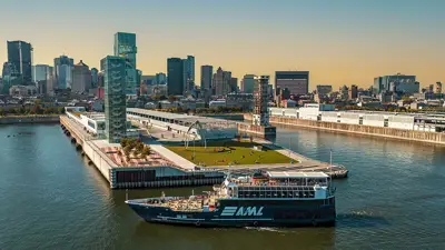 A blue cruise ship with the "AML" logo sails near a modern quay with green spaces, surrounded by the skyscrapers of a city under a clear and sunny sky.