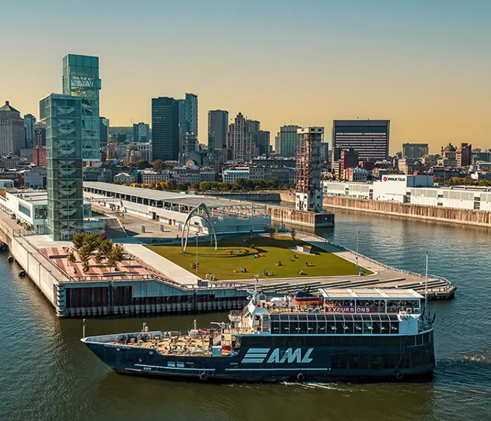 A blue cruise ship with the "AML" logo sails near a modern quay with green spaces, surrounded by the skyscrapers of a city under a clear and sunny sky.