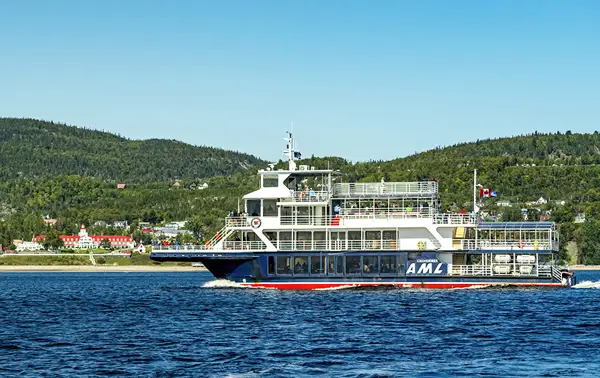 A large ferry with the letters "AML" sails on a body of water. It's a sunny day with clear skies. In the background, there are green hills and a small town with red-roofed buildings along the shoreline.
