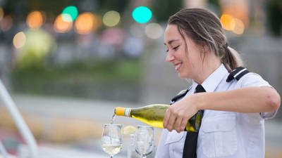 A smiling waitress in uniform pours white wine into glasses, with a blurred background of lights and greenery.