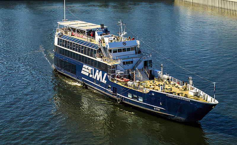 The ship "Cavalier Maxim" from AML cruises along a river towards an urban dock. The water is calm and slightly reflects the daylight. The boat is blue with white details.