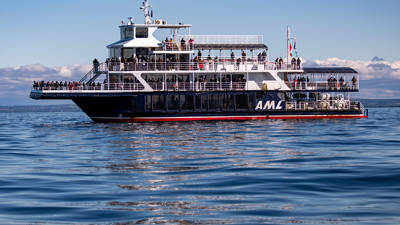 The ship "Le Grand Fleuve" from AML cruises sails during the day on calm waters, with passengers spread across multiple decks and a blue sky in the background.