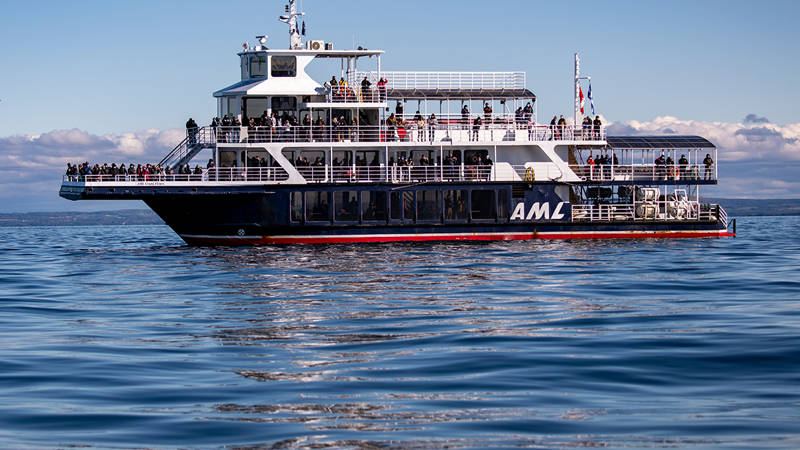 The ship "Le Grand Fleuve" from AML cruises sails during the day on calm waters, with passengers spread across multiple decks and a blue sky in the background.