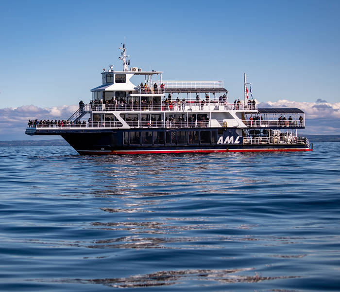 The ship "Le Grand Fleuve" from AML cruises sails during the day on calm waters, with passengers spread across multiple decks and a blue sky in the background.
