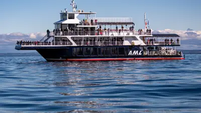 The ship "Le Grand Fleuve" from AML cruises sails during the day on calm waters, with passengers spread across multiple decks and a blue sky in the background.