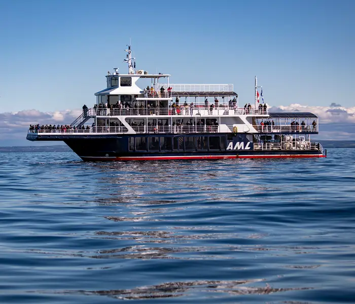 The ship "Le Grand Fleuve" from AML cruises sails during the day on calm waters, with passengers spread across multiple decks and a blue sky in the background.