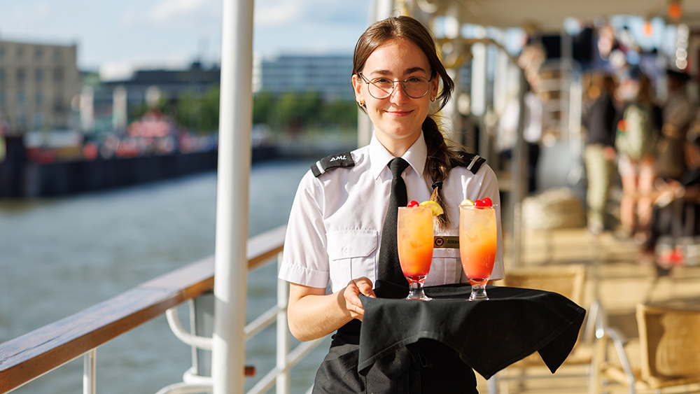 A waitress in uniform, wearing glasses and holding a tray with two colorful cocktails garnished with cherries. She stands on the deck of a boat, with a river and buildings in the background.