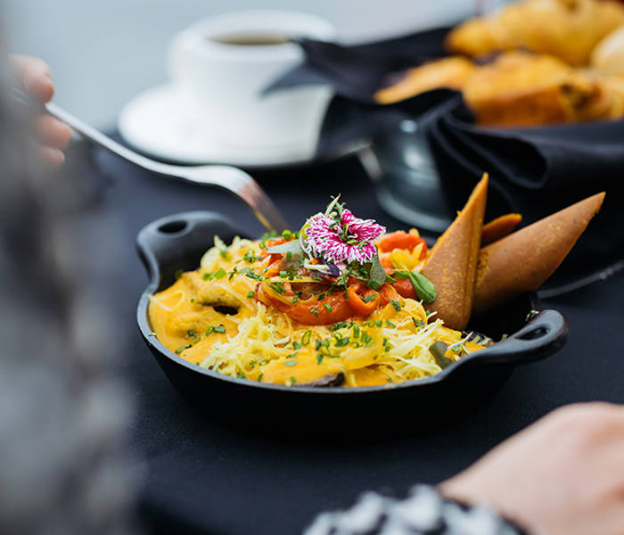 A colorful and elegant dish garnished with edible flowers, served in a small black skillet on a table with pastries, a cup of coffee, and a glass of juice.