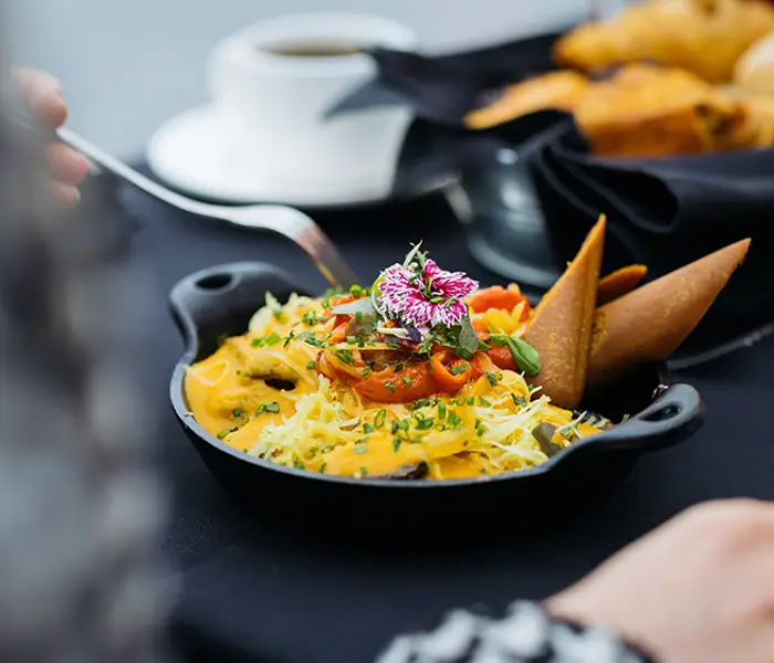 A colorful and elegant dish garnished with edible flowers, served in a small black skillet on a table with pastries, a cup of coffee, and a glass of juice.