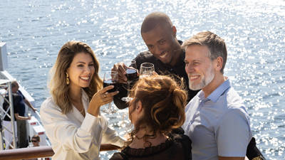 A group of smiling friends toasting with glasses of red wine on a boat deck, under bright sunlight, with a view of the water in the background.