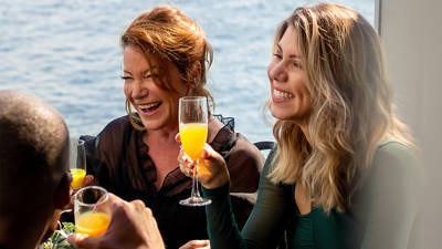 A group of friends enjoying a cheerful moment in a bright restaurant, with a smiling woman holding a glass of mimosa.
