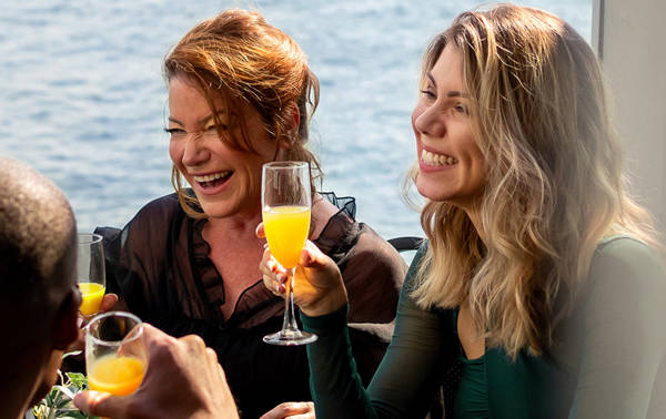 A group of friends enjoying a cheerful moment in a bright restaurant, with a smiling woman holding a glass of mimosa.