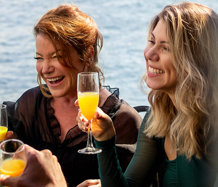 A group of friends enjoying a cheerful moment in a bright restaurant, with a smiling woman holding a glass of mimosa.