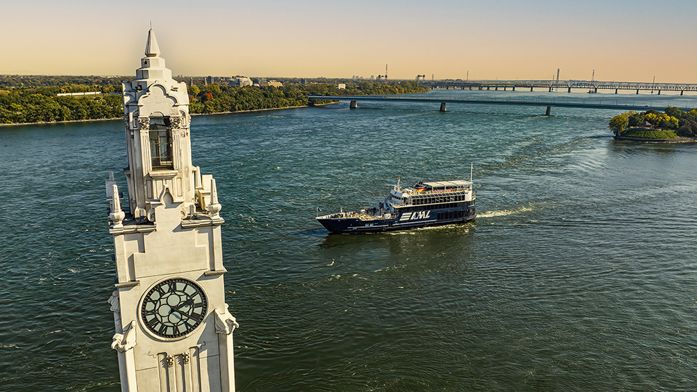 Aerial view of a tower with a large clock located by a river. A black cruise ship labeled "AML" sails on the water, with a bridge in the background and green riverbanks.