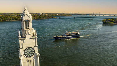 Aerial view of a tower with a large clock located by a river. A black cruise ship labeled "AML" sails on the water, with a bridge in the background and green riverbanks.