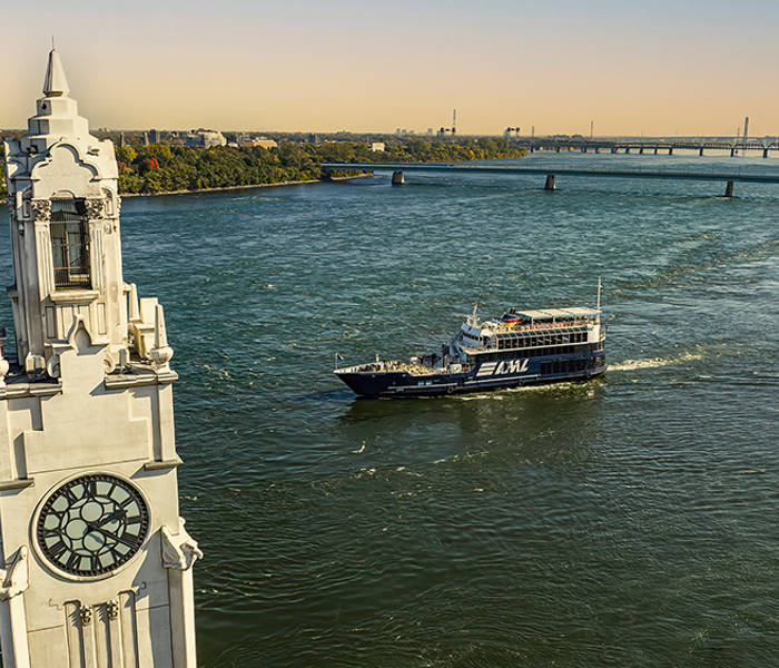 Aerial view of a tower with a large clock located by a river. A black cruise ship labeled "AML" sails on the water, with a bridge in the background and green riverbanks.