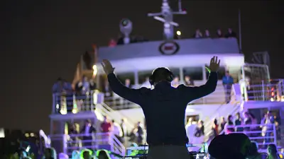 Un DJ anime une soirée à bord d'un bateau, les bras levés, face à une foule de passagers sur les ponts supérieurs. Des lumières colorées illuminent la scène, créant une ambiance festive sous le ciel nocturne.