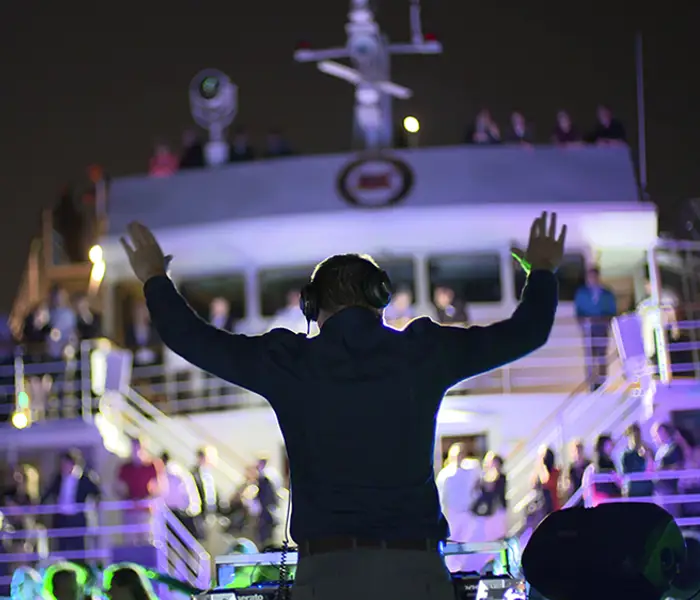 Un DJ anime une soirée à bord d'un bateau, les bras levés, face à une foule de passagers sur les ponts supérieurs. Des lumières colorées illuminent la scène, créant une ambiance festive sous le ciel nocturne.