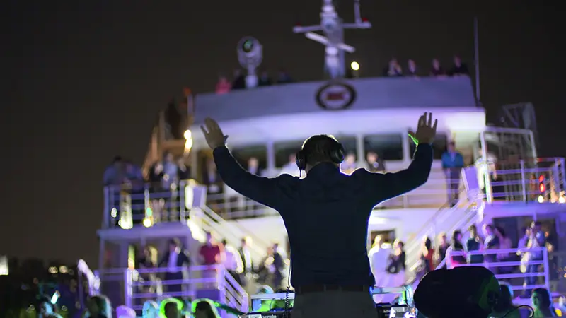 Un DJ anime une soirée à bord d'un bateau, les bras levés, face à une foule de passagers sur les ponts supérieurs. Des lumières colorées illuminent la scène, créant une ambiance festive sous le ciel nocturne.