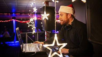  A DJ wearing a Santa hat mixes music on his laptop and turntables in a festive setting, surrounded by star-shaped light decorations.
