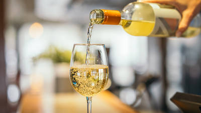 A close-up of a hand pouring white wine from a bottle into a wine glass. The wine is bubbly and fills the glass on a blurred background, creating a warm and inviting atmosphere.