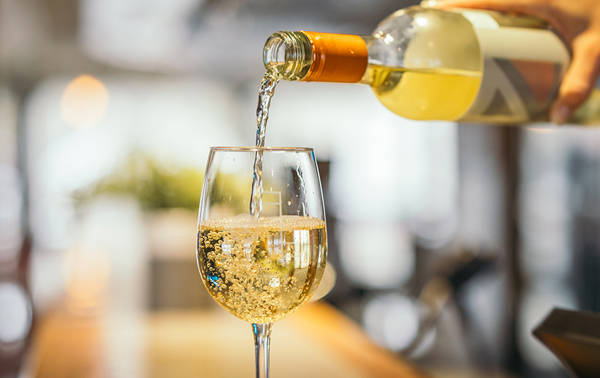 A close-up of a hand pouring white wine from a bottle into a wine glass. The wine is bubbly and fills the glass on a blurred background, creating a warm and inviting atmosphere.