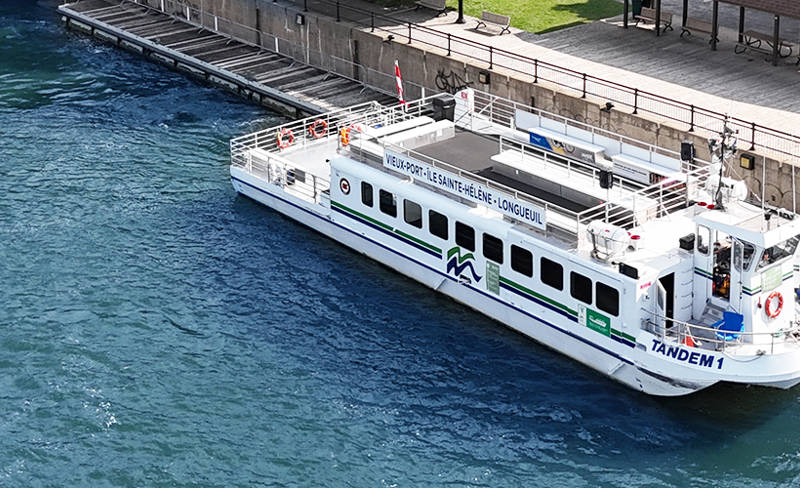 Le bateau "Tandem" est amarré le long d'un quai bordé de verdure. On aperçoit des arbres, un chemin piétonnier, des bâtiments avec des toits en pente, et un parking à proximité. L'eau est calme, et la scène se déroule par une journée ensoleillée.