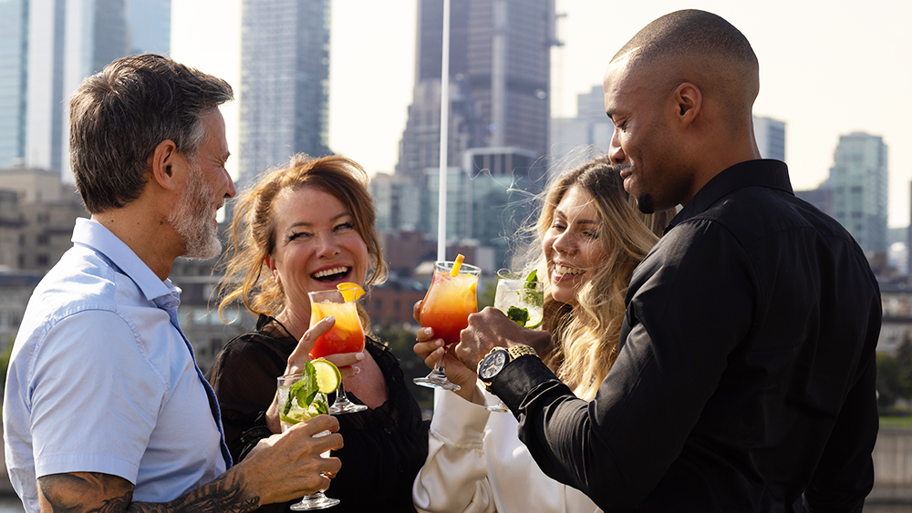 A group of four smiling friends toasting with colorful cocktails on a terrace, with skyscrapers in the background.