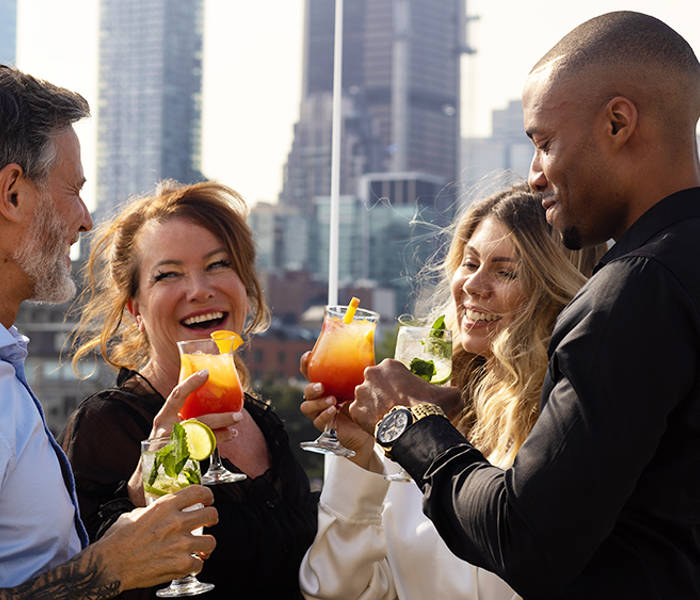A group of four smiling friends toasting with colorful cocktails on a terrace, with skyscrapers in the background.