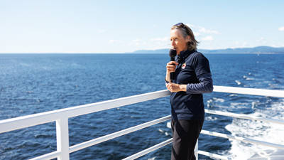 A naturalist holds a microphone and gives a presentation on the deck of a boat, with a clear view of the blue ocean in the background. She is wearing a navy-blue sweater and stands near the white railing under a sunny, clear sky, sharing information about the surrounding nature.