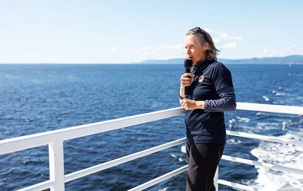 A naturalist holds a microphone and gives a presentation on the deck of a boat, with a clear view of the blue ocean in the background. She is wearing a navy-blue sweater and stands near the white railing under a sunny, clear sky, sharing information about the surrounding nature.