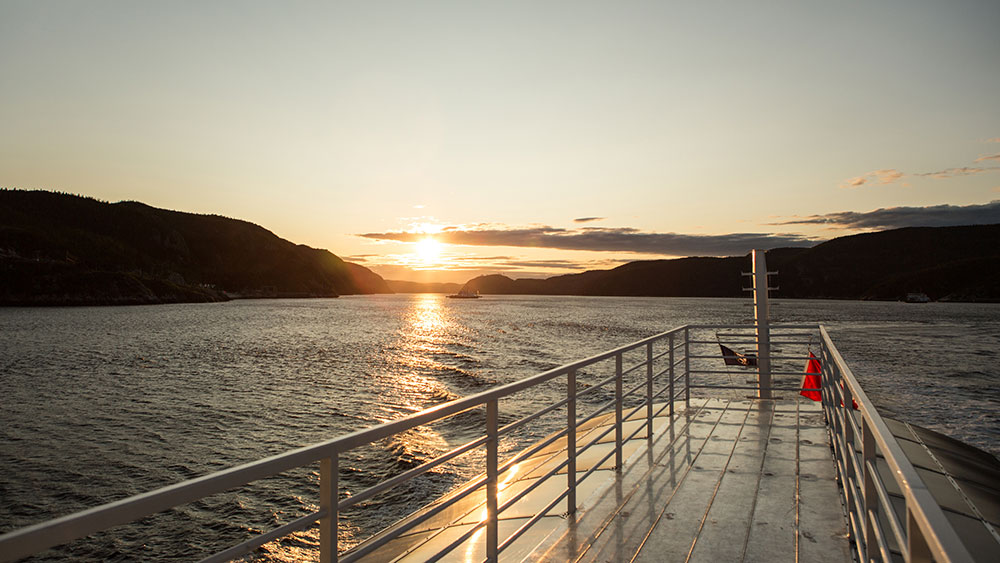 View from the deck of a boat at sunset, with a river bordered by dark hills on the horizon. The golden sunlight reflects on the water and the shiny metal deck, creating a peaceful and warm atmosphere.