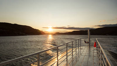 View from the deck of a boat at sunset, with a river bordered by dark hills on the horizon. The golden sunlight reflects on the water and the shiny metal deck, creating a peaceful and warm atmosphere.