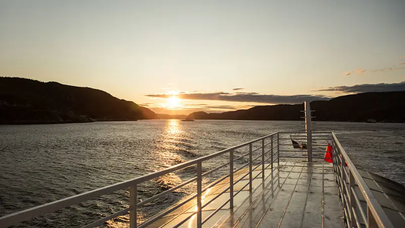 Vue depuis le pont d'un bateau au coucher du soleil, avec une rivière bordée de collines sombres à l'horizon. La lumière dorée du soleil se réfléchit sur l'eau et sur le métal brillant du pont, créant une ambiance paisible et chaleureuse.