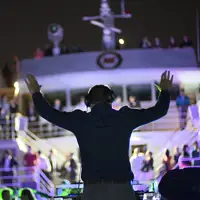 Un DJ anime une fête nocturne sur un bateau, avec des lumières colorées et une foule dansante sur plusieurs niveaux du navire.